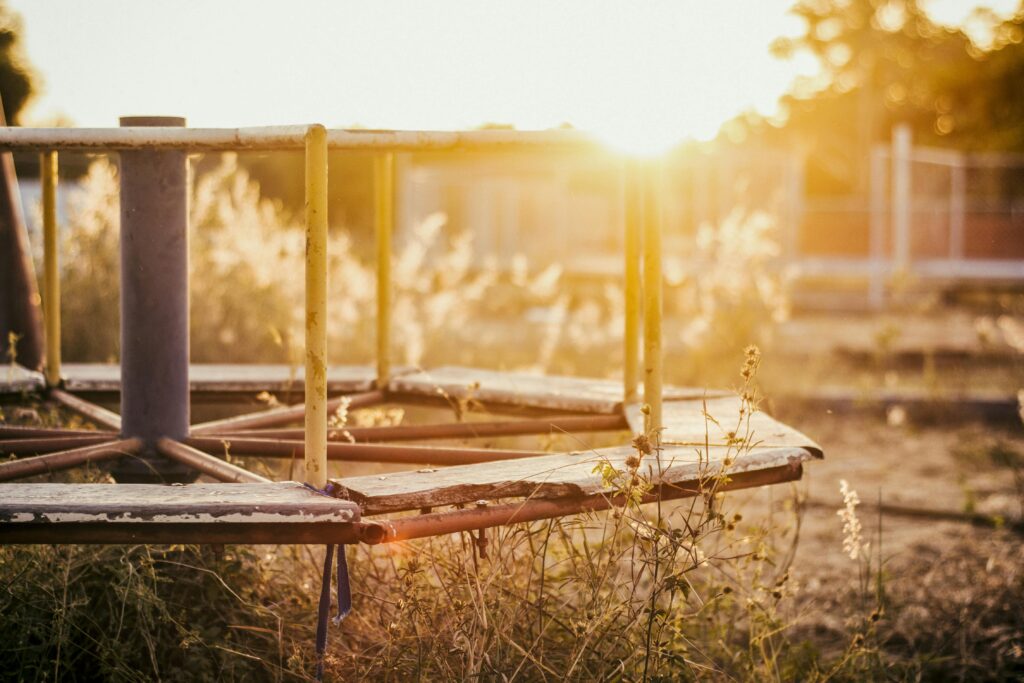 A sunlit, rusted merry-go-round in an abandoned playground overgrown with grass.